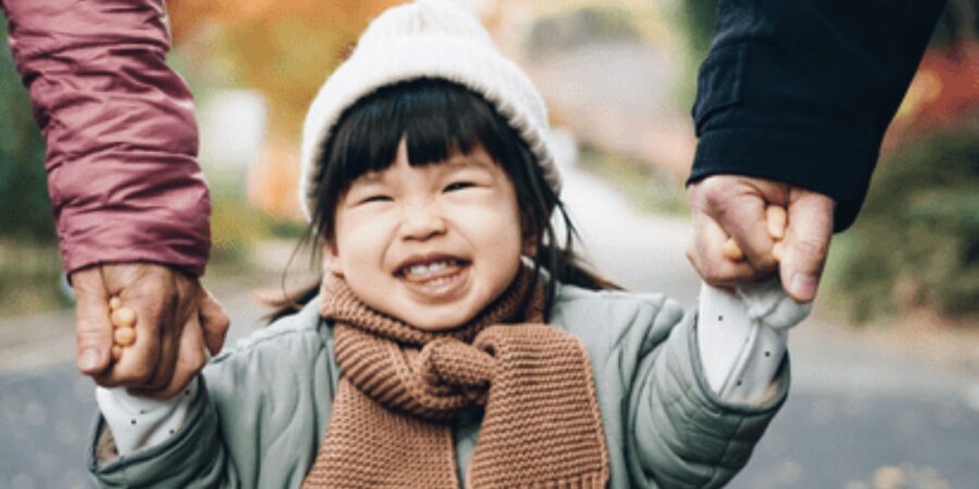 Smiling child holding adults' hands outdoors.